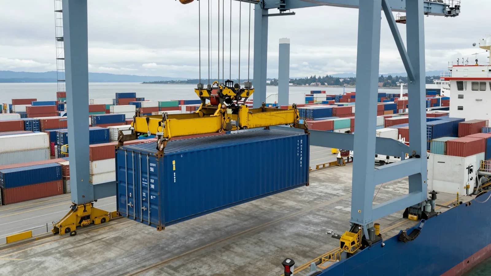 Grúa portuaria moviendo un contenedor azul sobre una terminal llena de contenedores.