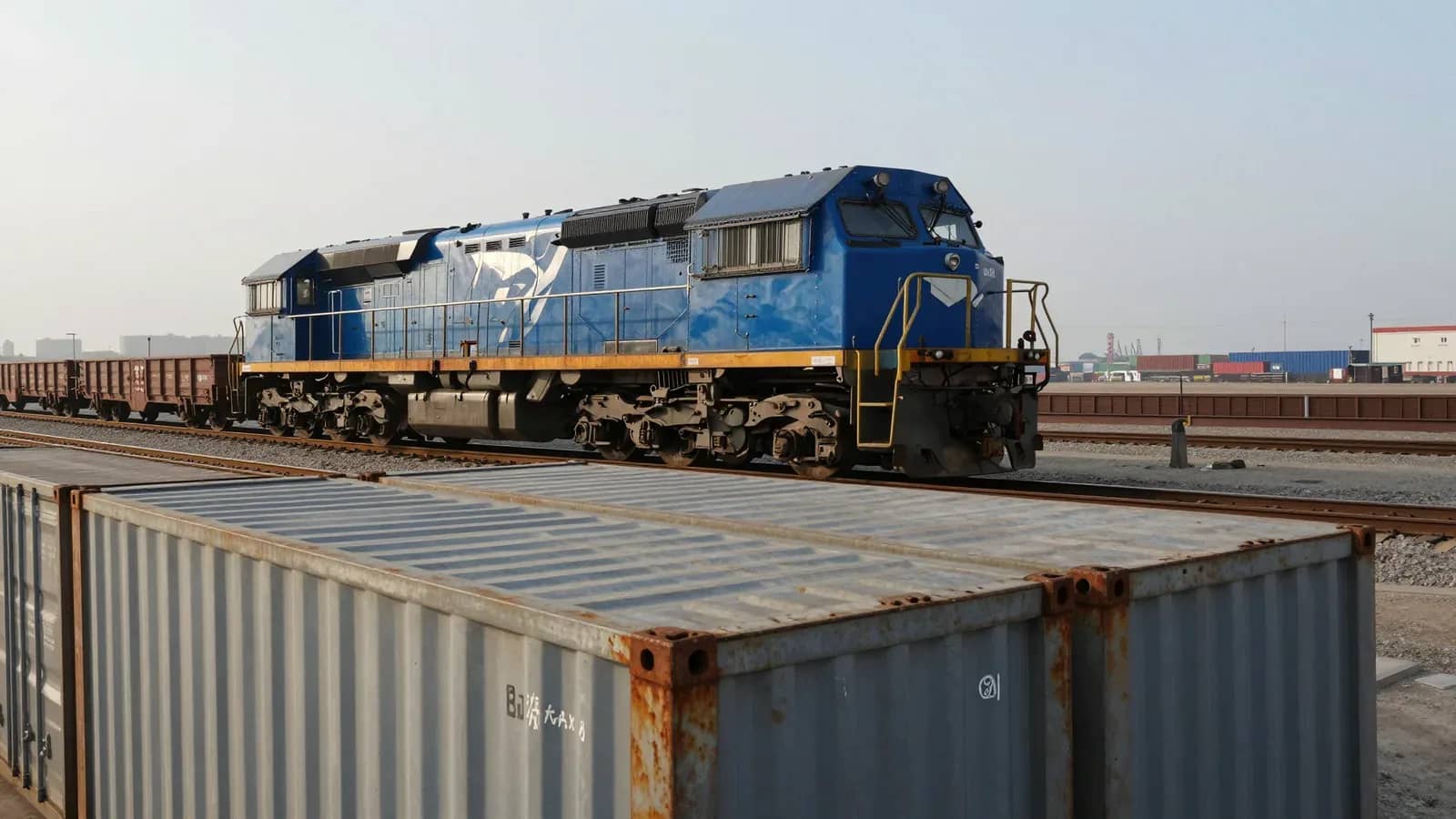 Locomotora azul y vagones de carga en un patio ferroviario junto a contenedores portuarios