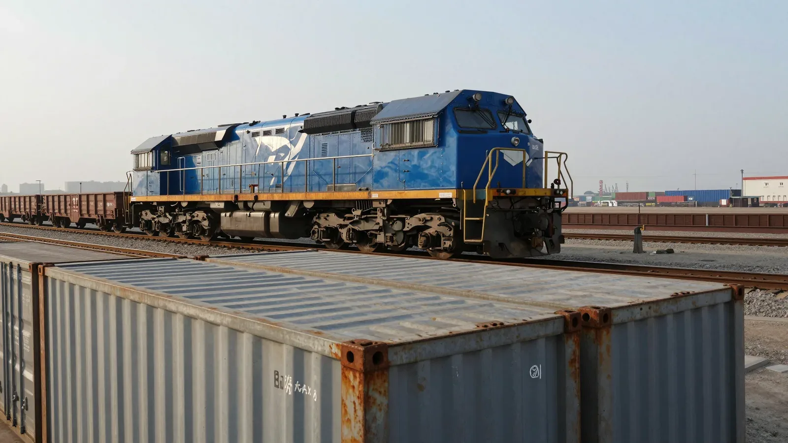 Locomotora azul y vagones de carga en un patio ferroviario junto a contenedores portuarios