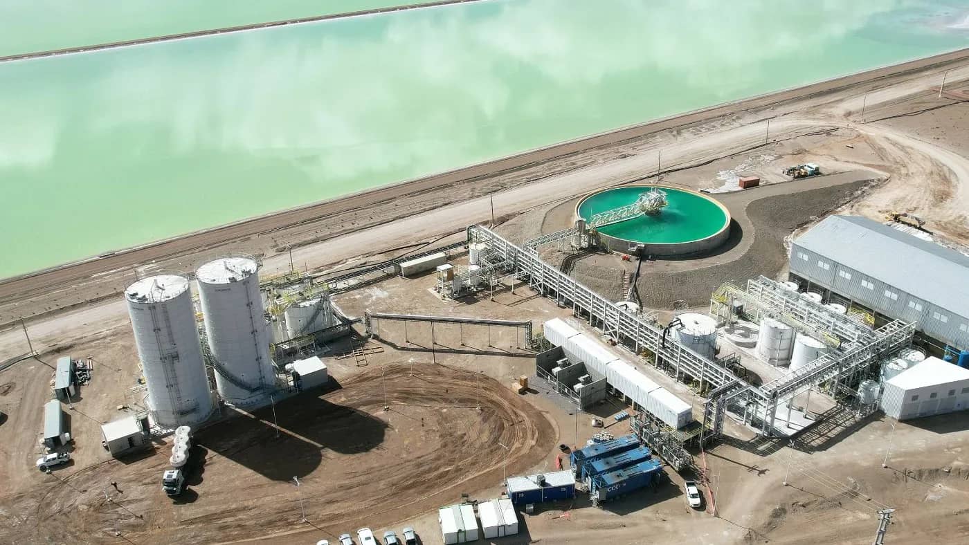 Vista aérea de planta de procesamiento de litio con estanques de evaporación turquesas, tanques de almacenamiento cilíndricos blancos, estructuras industriales y vehículos en terre